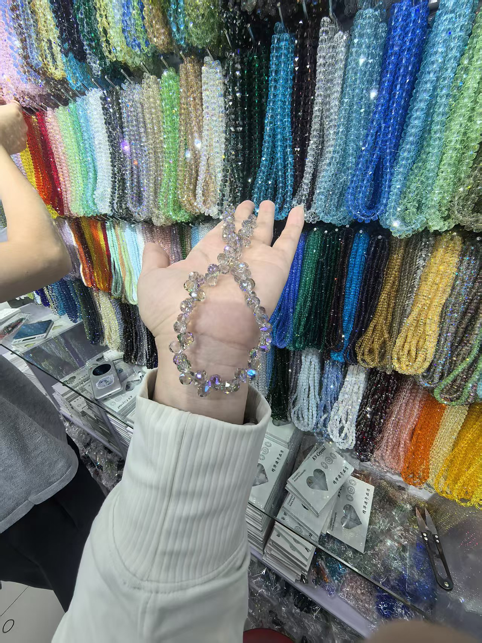 Hand holding gray faceted crystal beads in front of a multicolor wholesale bead wall